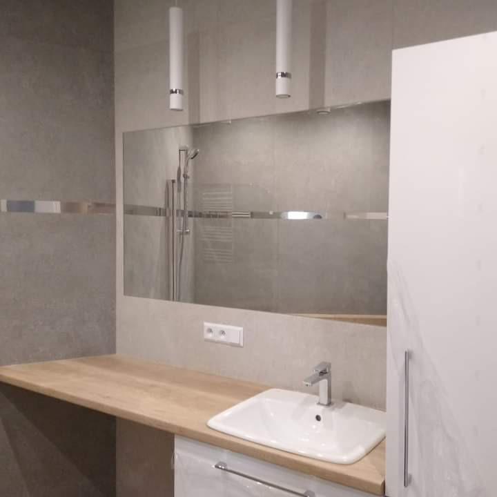 Jasna łazienka po renowacji: umywalka wpuszczona w drewniany blat, duże lustro nad blatem, widoczna kabina prysznicowa, nowoczesne oświetlenie punktowe.