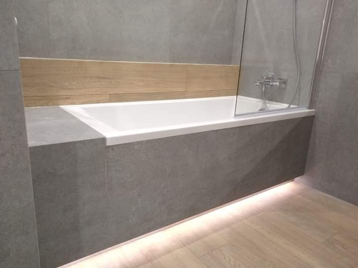 Nowoczesna łazienka z wanną obudowaną szarymi płytkami, drewnianym akcentem nad wanną i subtelnym podświetleniem LED u dołu, oddzielona szklanym parawanem.