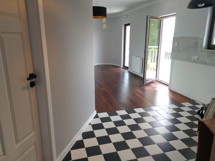 Wnętrze mieszkania po remoncie: szaro-białe ściany, ciemna podłoga z paneli, czarno-biała podłoga w szachownicę przy wejściu, białe drzwi z czarną klamką i widok na balkon.