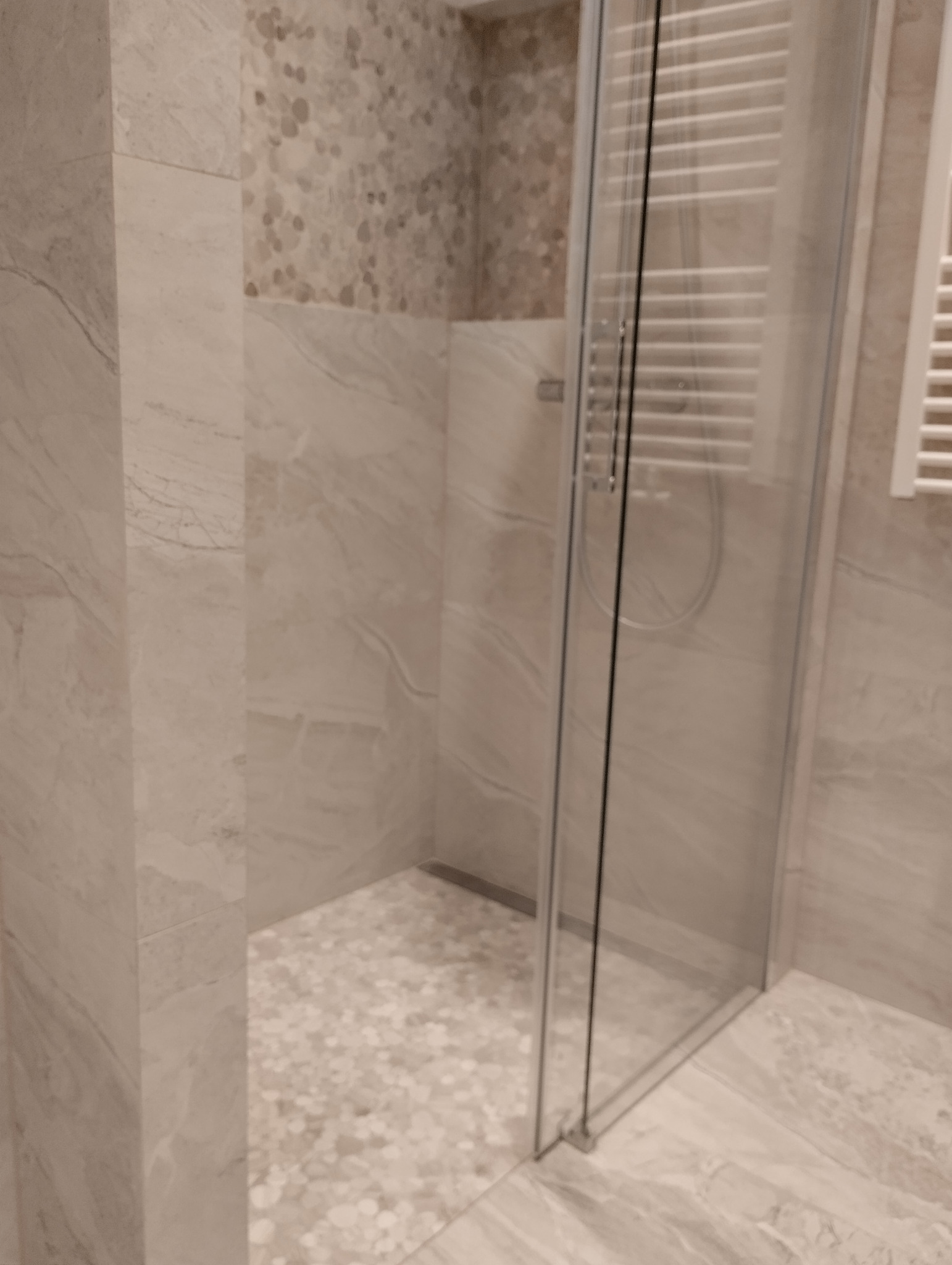 Beżowa łazienka z kabiną prysznicową typu walk-in, wykończona płytkami imitującymi marmur i mozaiką na podłodze strefy prysznicowej, z grzejnikiem drabinkowym w tle.