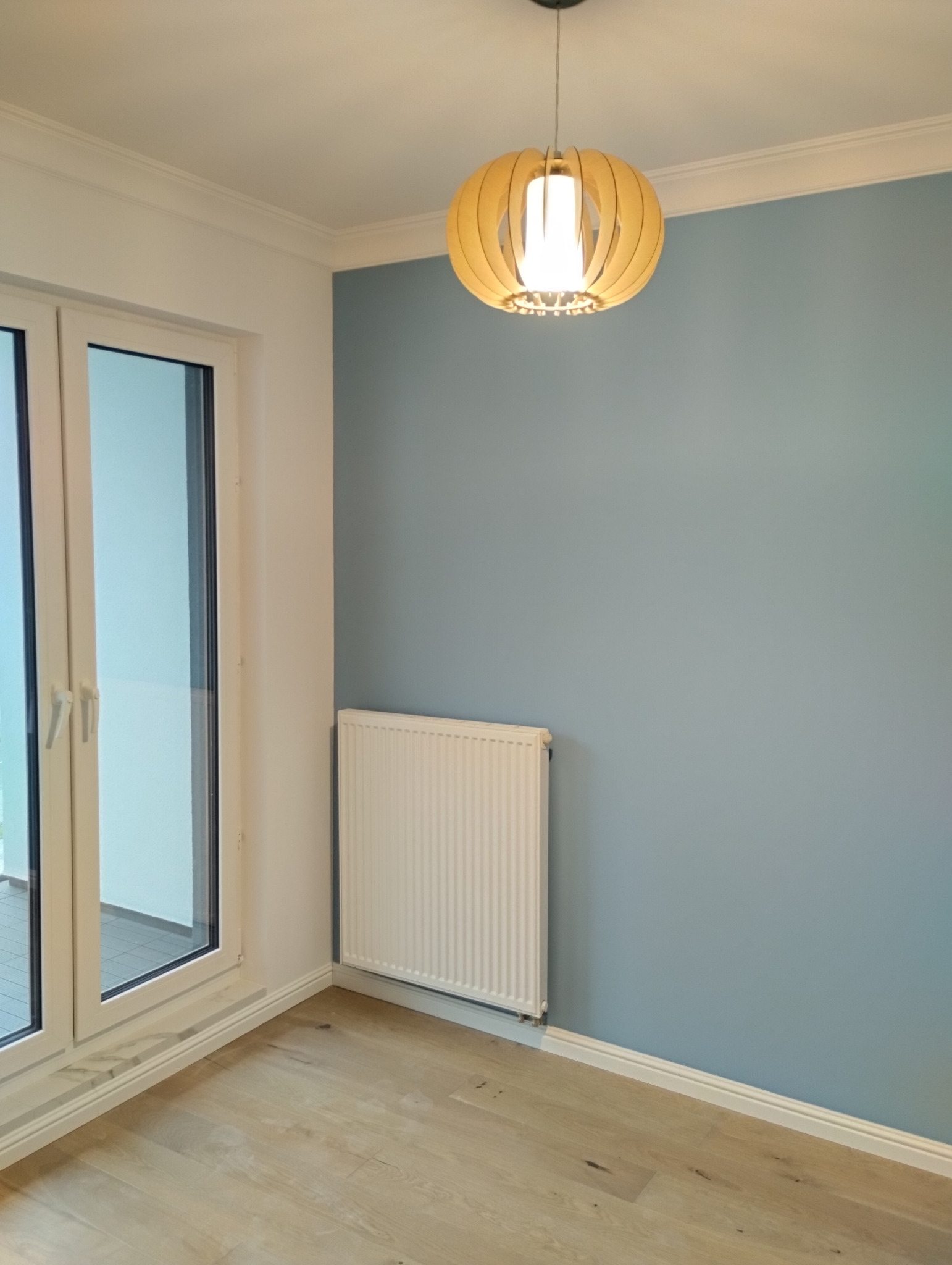 Świeżo pomalowane na błękitno wnętrze pokoju w Zielonce z drewnianą podłogą i białym grzejnikiem pod ścianą, oświetlone nowoczesną lampą.