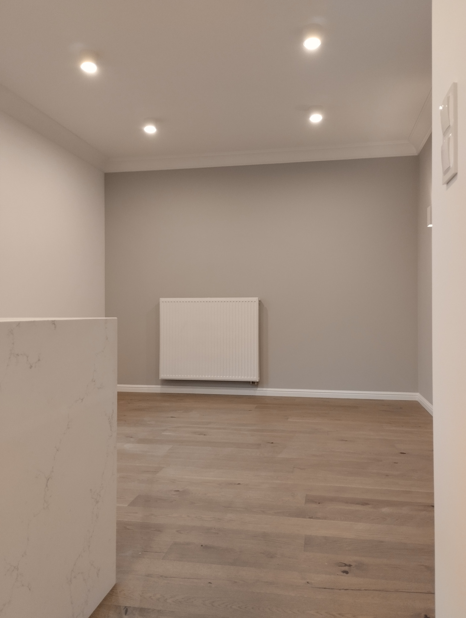 Pomalowane na szaro wnętrze z drewnianą podłogą, białym grzejnikiem i czterema punktowymi światłami w suficie, fragment blatu z marmurowym wzorem po lewej stronie
