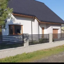 Nowoczesne ogrodzenie panelowe z poziomych profili w kolorze czarnym, zamontowane na betonowych słupkach i podmur&oacute;wce, na tle białego domu z ciemnym dachem.