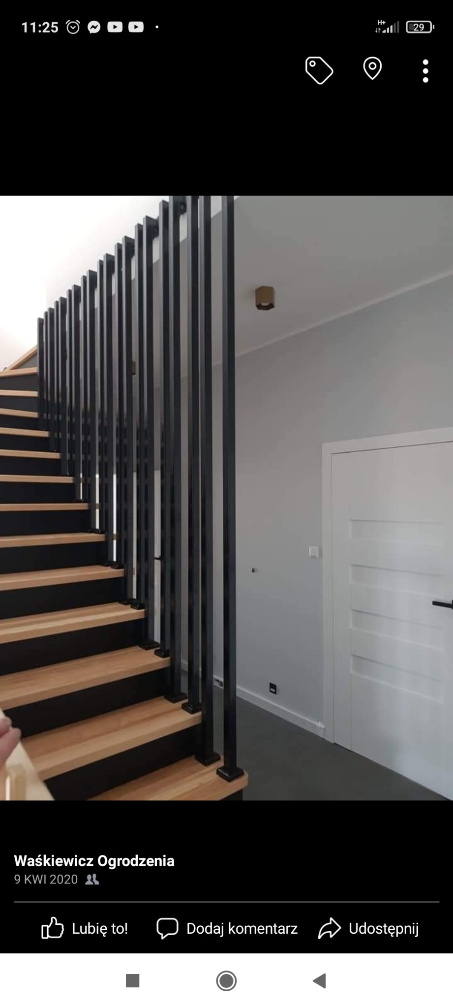 Nowoczesne schody drewniane z czarnymi, metalowymi balustradami pionowymi, prowadzące do białych drzwi w szarym pomieszczeniu.