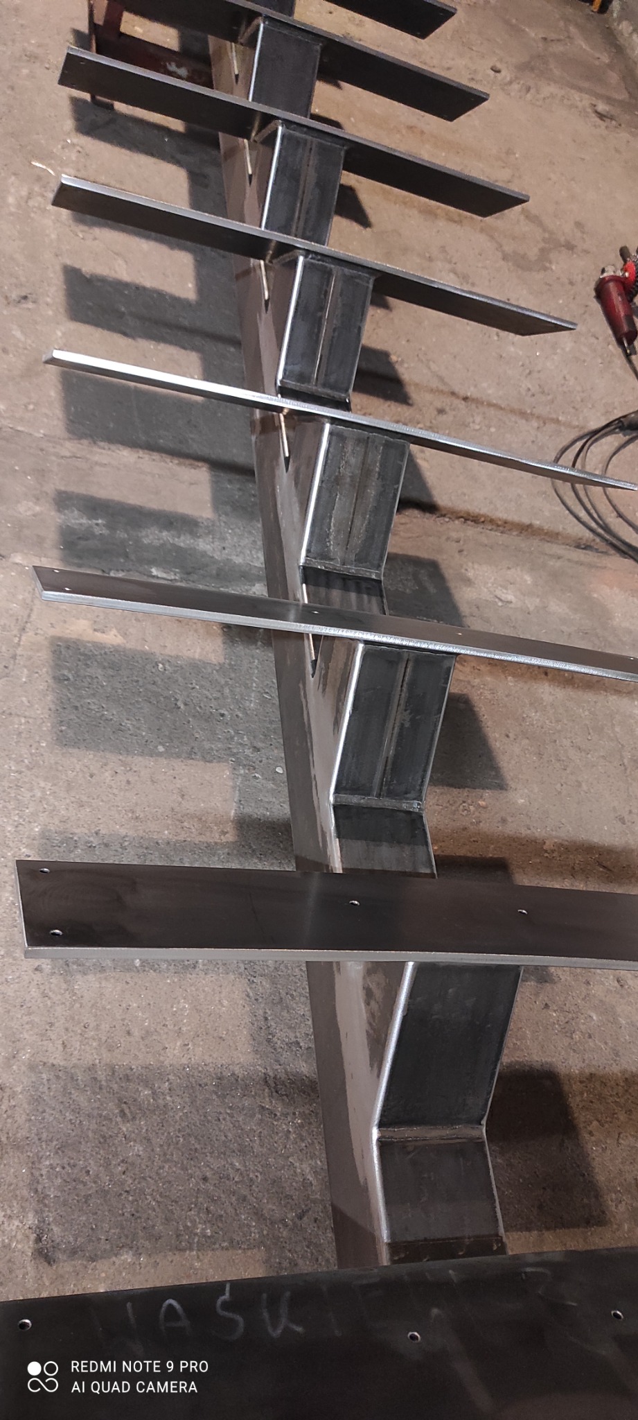 Surowa, metalowa konstrukcja schodów z przyspawanymi stopniami, widoczne spawy i otwory montażowe, w tle szlifierka kątowa.