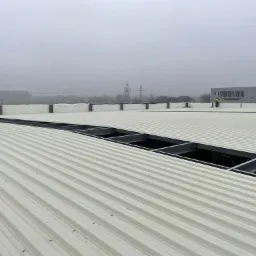 Montaż blachy trapezowej na dachu hali przemysłowej w pochmurny dzień, widoczne stalowe profile konstrukcyjne pod blachą oraz pracownik w odblaskowej kamizelce w tle, w oddali budynek z szyldem.