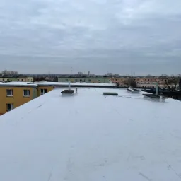 Widok z góry na biały, płaski dach budynku wielorodzinnego z kominami wentylacyjnymi na tle zabudowy miejskiej i pochmurnego nieba.