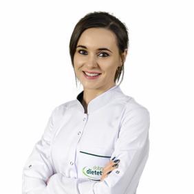 Uśmiechnięta kobieta w białym fartuchu medycznym z logo 'dobry dietetyk' na kieszeni, pozująca na jasnym tle z założonymi rękami.