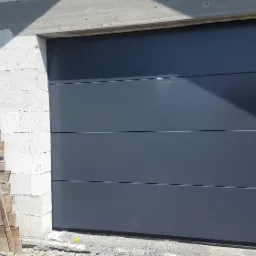 Zewnętrzna, segmentowa brama garażowa w kolorze grafitowym zamontowana w surowym otworze budynku z betonu komórkowego, obok leżą drewniane deski szalunkowe.