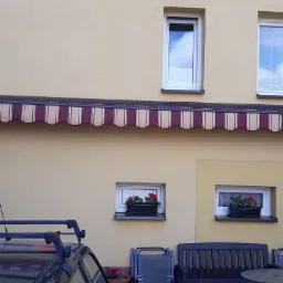 Zewnętrzna markiza w paski, zamontowana nad oknami na elewacji budynku z jasną fasadą. Widoczne także rośliny w doniczkach na parapetach i fragment samochodu z bagażnikiem dachowym.