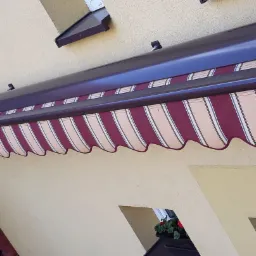 Zewnętrzna markiza w burgundowo-kremowe pasy, zamontowana nad oknem, z widocznym fragmentem elewacji budynku i rynną w kolorze markizy.