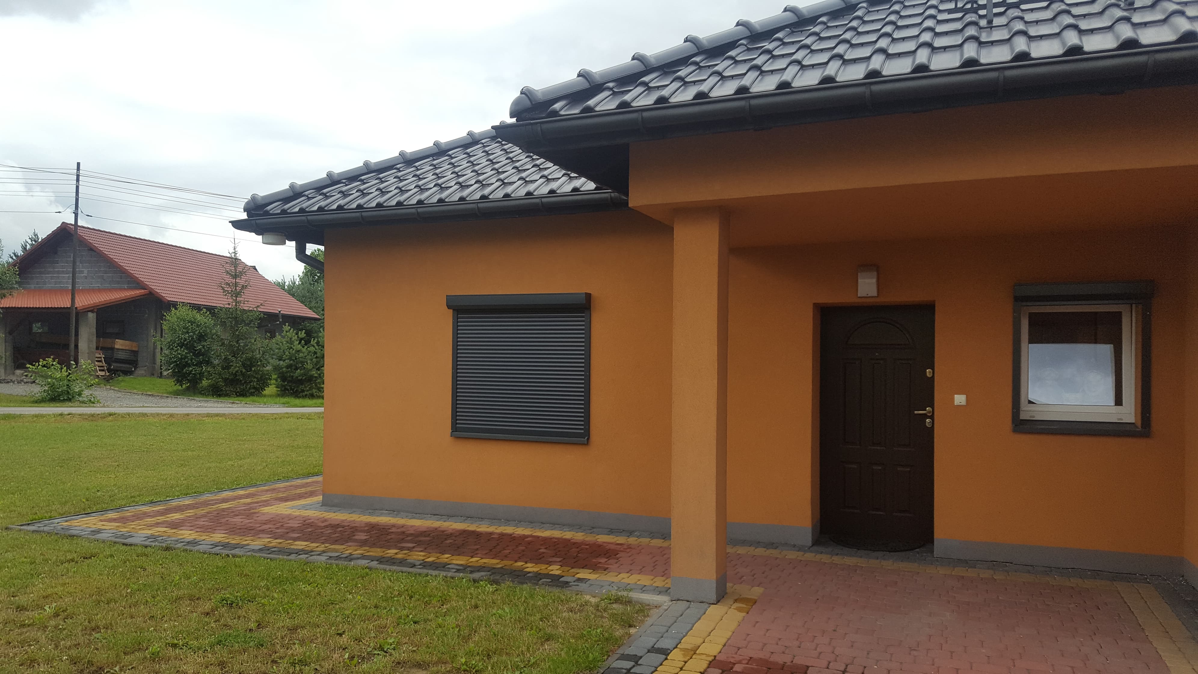 Dom z pomarańczową elewacją, grafitowym dachem, zamkniętą szarą roletą zewnętrzną na oknie i ciemnymi drzwiami wejściowymi, widoczny z zewnątrz na tle zielonego trawnika i drugiego budynku.