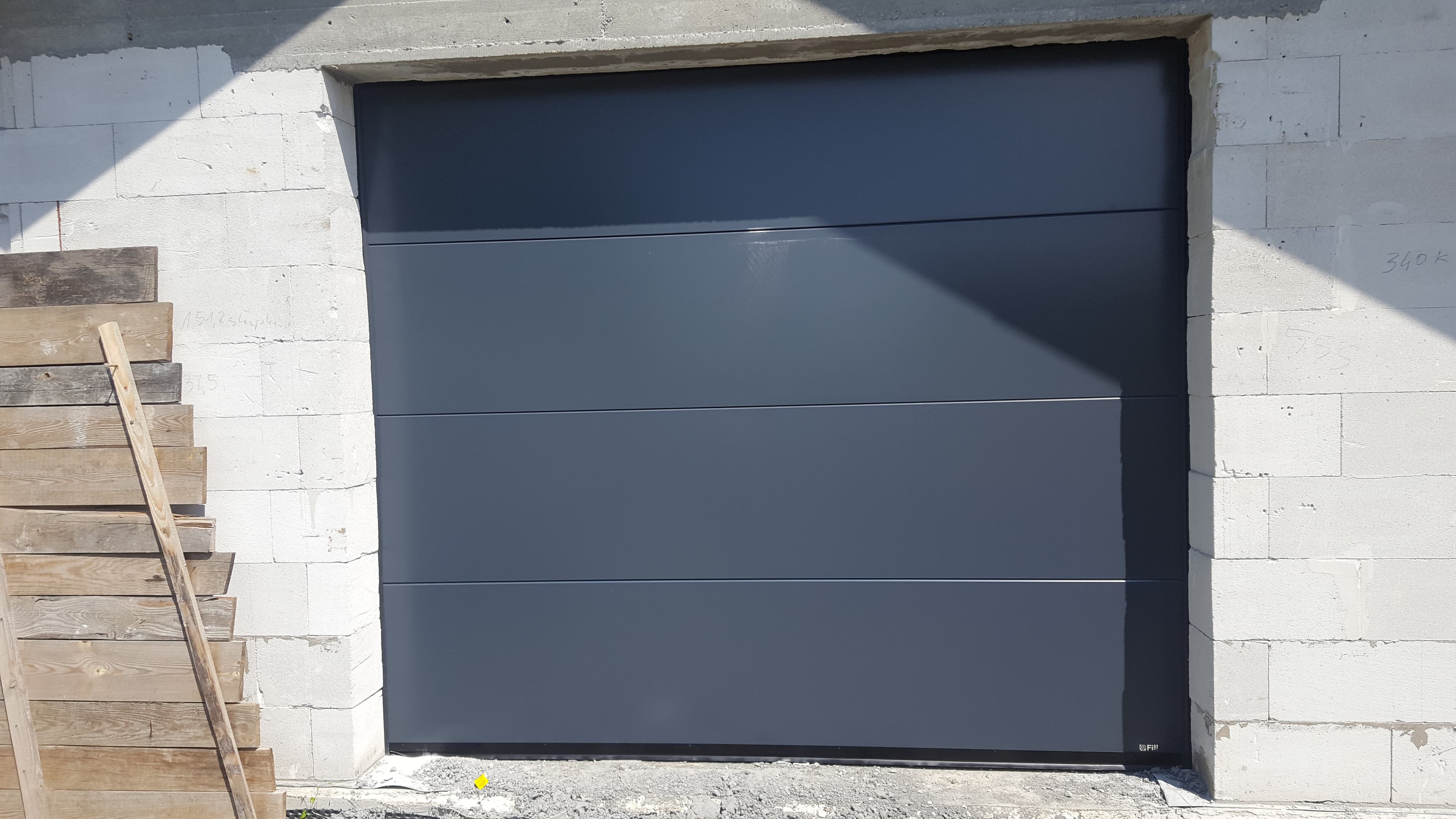 Szara, segmentowa brama garażowa w surowym otworze budynku z betonu komórkowego, widoczne drewniane podpory i niedokończone podłoże.