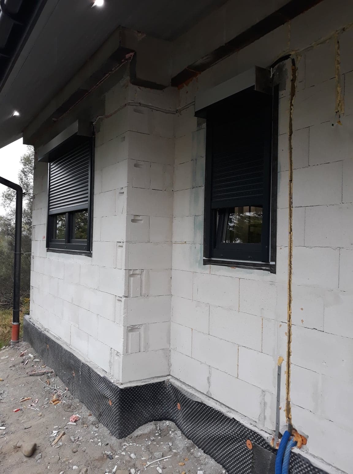 Dwa okna z czarnymi roletami zewnętrznymi zamontowane w surowej ścianie z białych bloczków, widoczne instalacje elektryczne i rury na zewnątrz budynku.