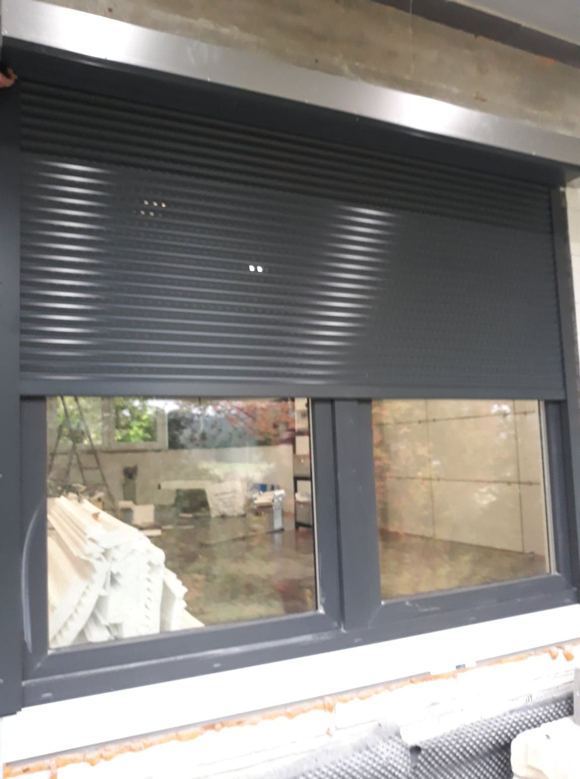 Grafitowa roleta zewnętrzna częściowo opuszczona na oknie PCV, widoczne wnętrze w trakcie remontu z materiałami budowlanymi i drabiną.