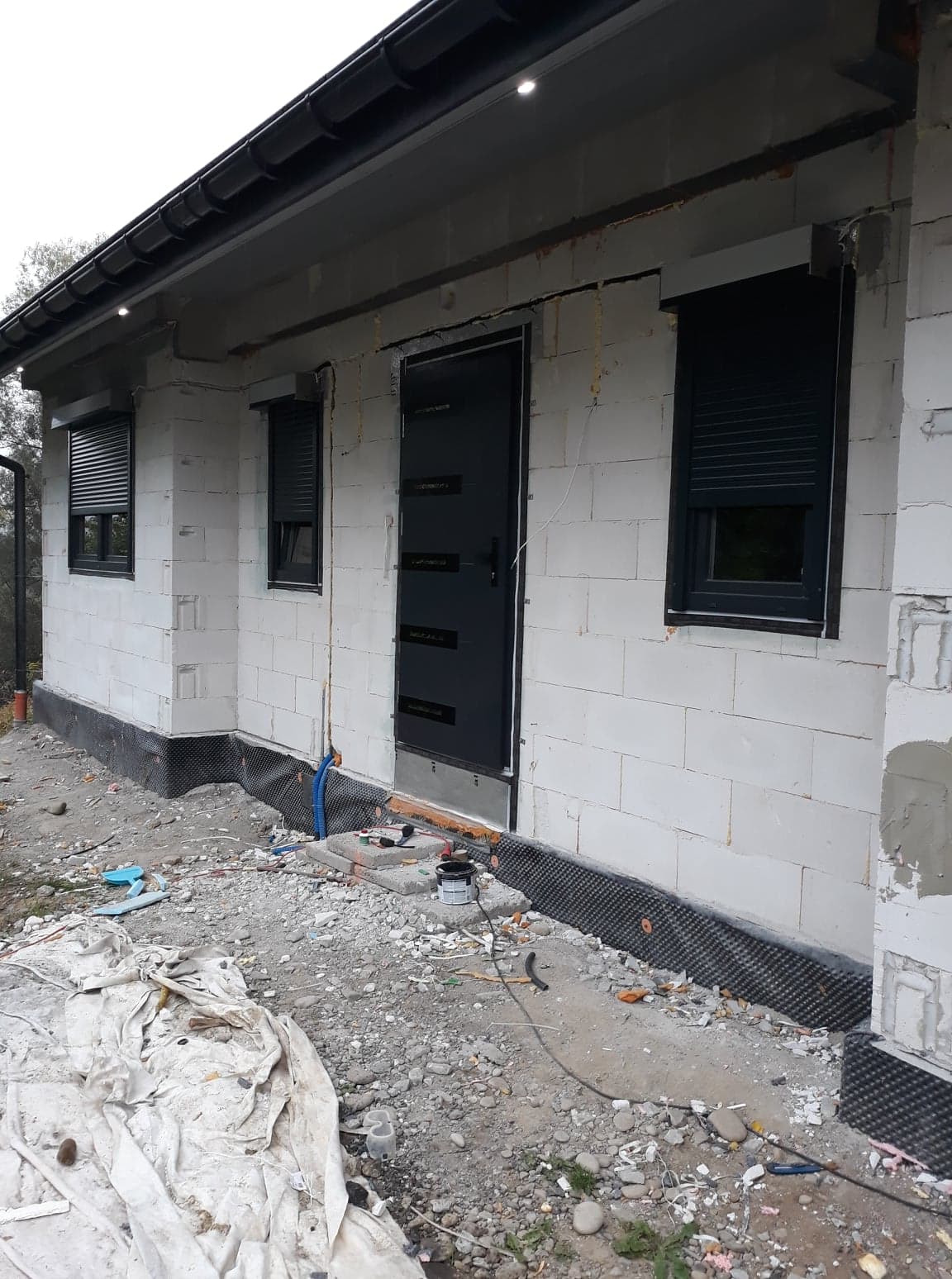 Surowy stan elewacji domu jednorodzinnego z zamontowanymi oknami z roletami zewnętrznymi i czarnymi drzwiami wejściowymi, widoczne ślady prac wykończeniowych na zewnątrz budynku.