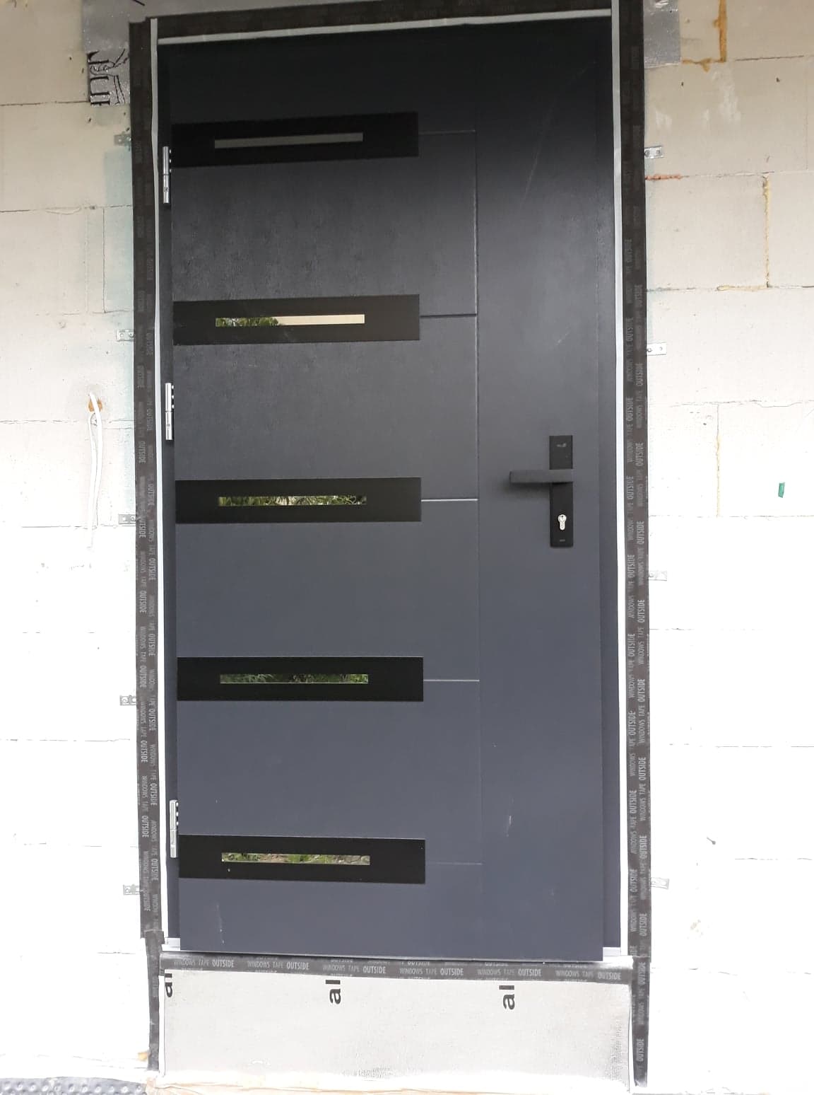 Szare drzwi wejściowe z poziomymi przeszkleniami, zamontowane w surowym murze z białych bloczków, zabezpieczone taśmą montażową.