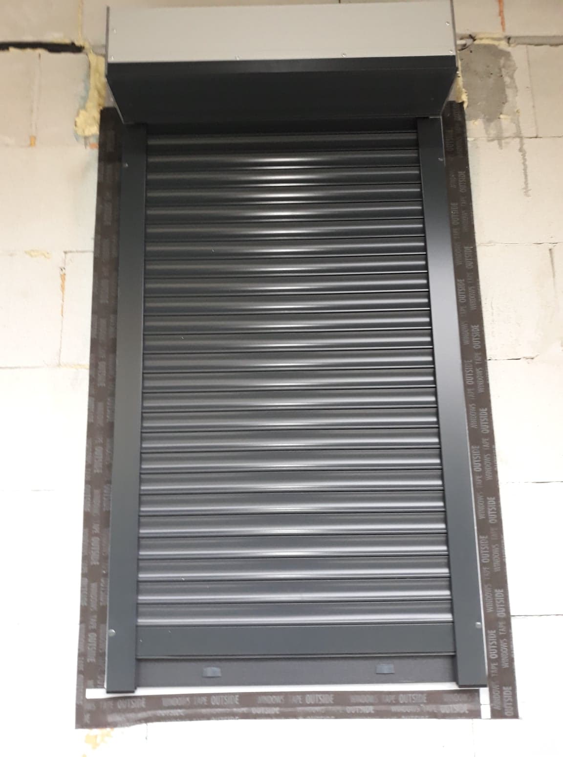 Zamknięta, szara roleta zewnętrzna zamontowana na oknie w Wadowicach, widoczna taśma uszczelniająca na ramie okiennej, surowy mur w tle.