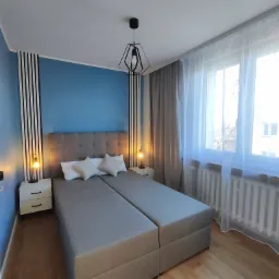 Nowoczesna sypialnia z dwoma połączonymi łóżkami, niebieską ścianą z dekoracyjnymi czarno-białymi pasami, białymi szafkami nocnymi z lampkami, geometryczną lampą wiszącą i oknem z białymi firankami.