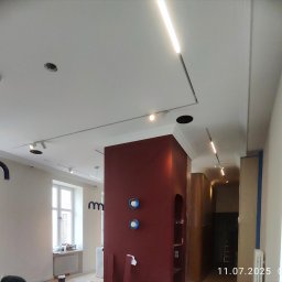 PAB PATRYK BIEŃ - Wnętrze w trakcie wykańczania: biały sufit z nowoczesnym oświetleniem LED, bordowa ściana z elementami dekoracyjnymi, okna z grzejnikami. Widoczne prace wykończeniowe.