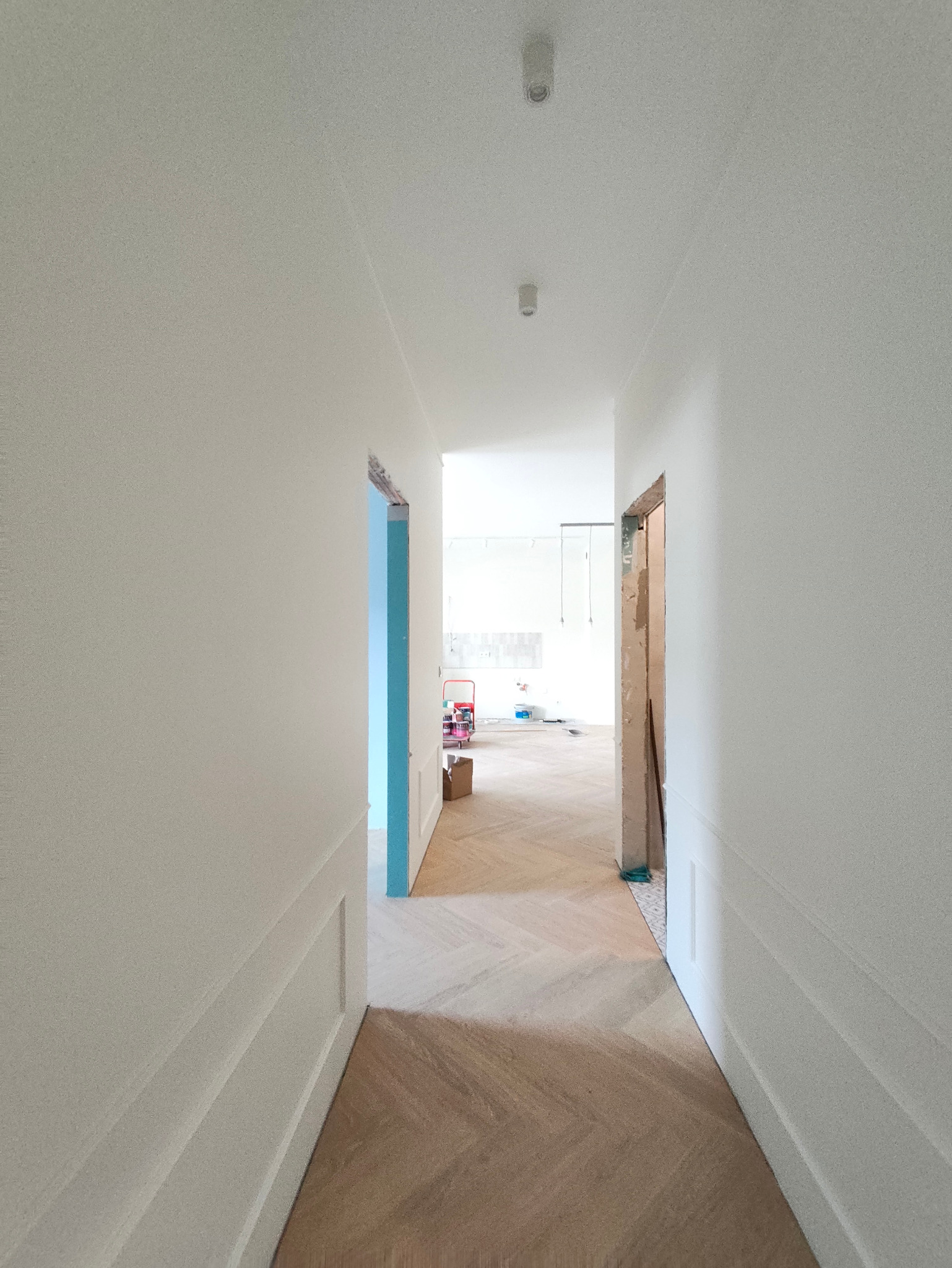 Korytarz w trakcie remontu z parkietem w jodełkę, świeżo pomalowanymi ścianami i widocznymi otworami drzwiowymi. W tle widoczny wózek malarski.