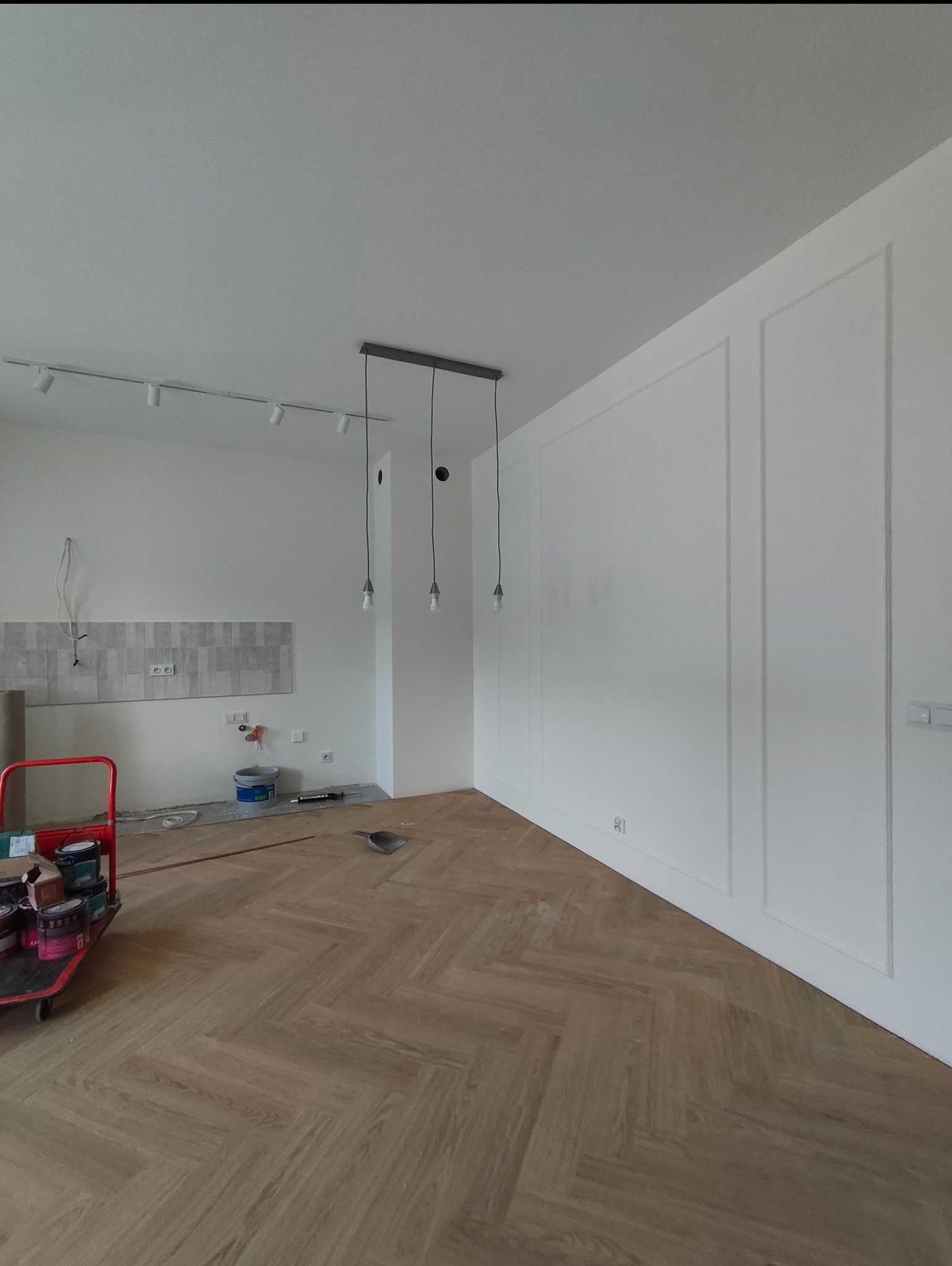 Wnętrze w trakcie wykańczania: parkiet w jodełkę, białe ściany z dekoracyjnymi listwami, sufit z oświetleniem szynowym i wiszącymi lampami. W tle płytki i wózek z farbami.
