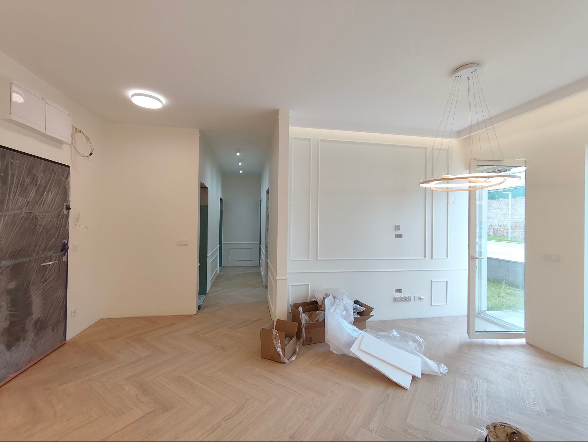 Jasne wnętrze po remoncie z parkietem w jodełkę, ozdobnymi listwami na ścianach i nowoczesną lampą. Częściowo widoczne materiały budowlane.