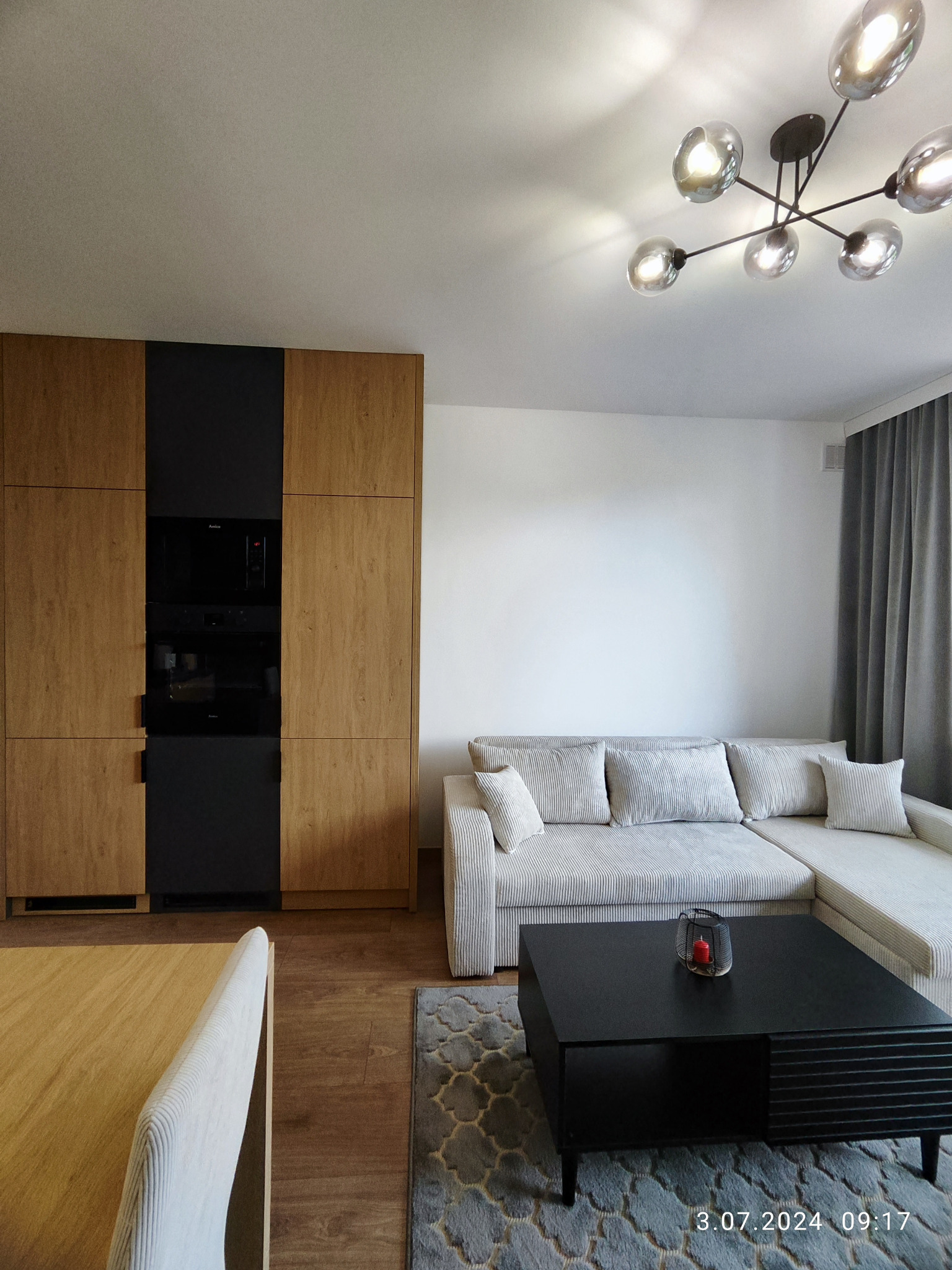 Nowoczesny salon z zabudową kuchenną w odcieniach drewna i czerni, szary narożnik i dywan. Oświetlenie sufitowe w stylu industrialnym. Minimalistyczny design.