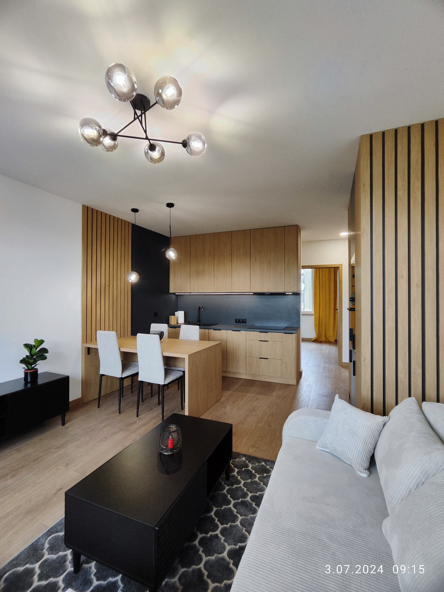 Nowoczesne wnętrze: drewniana zabudowa kuchni, stół z krzesłami, czarny stolik kawowy na wzorzystym dywanie, designerska lampa sufitowa i dekoracyjne lamele ścienne.