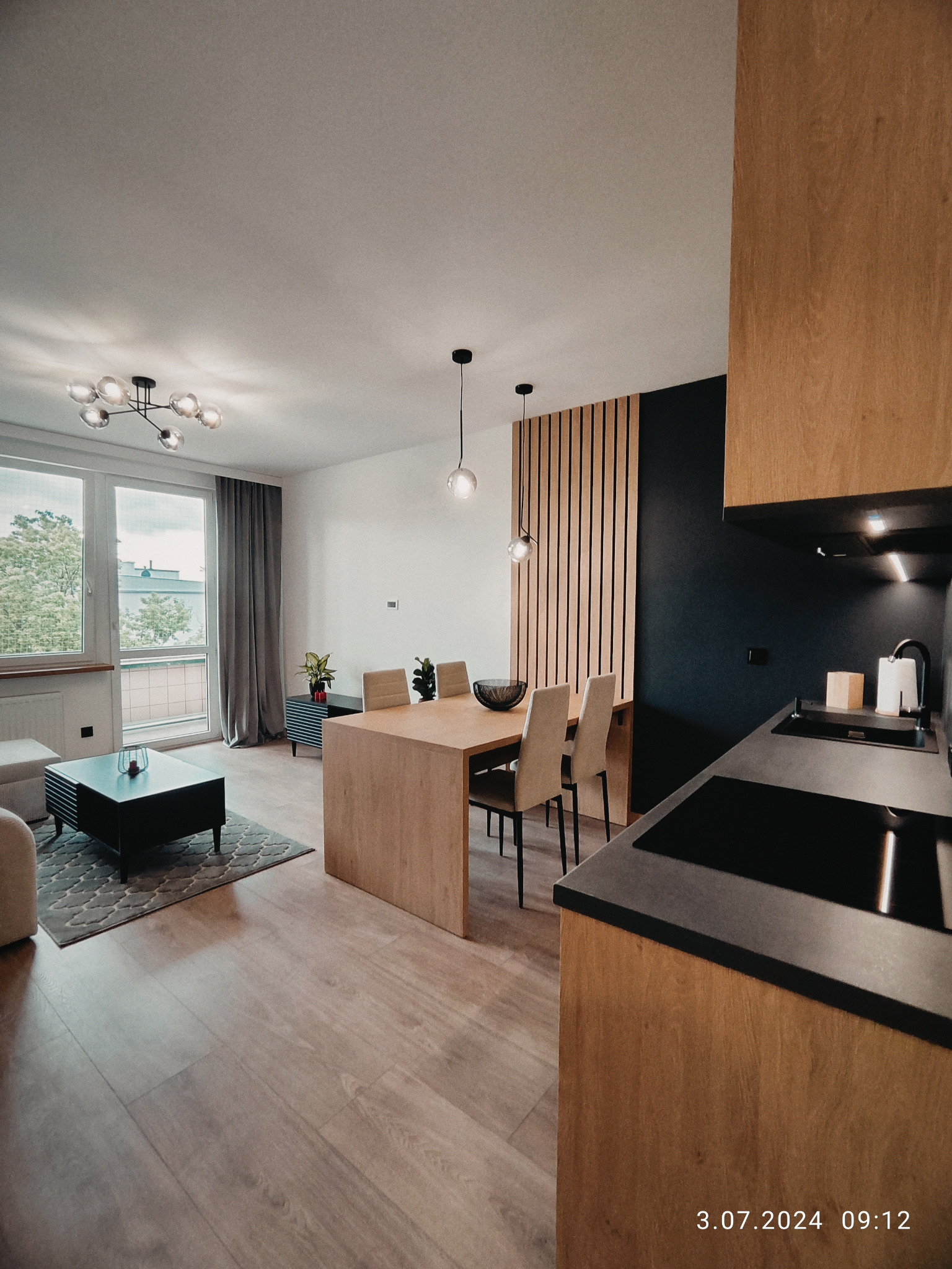 Nowoczesne wnętrze mieszkania z drewnianą zabudową kuchenną, stołem jadalnym i elementami dekoracyjnymi. Widok na salon z oknem i minimalistycznym oświetleniem.