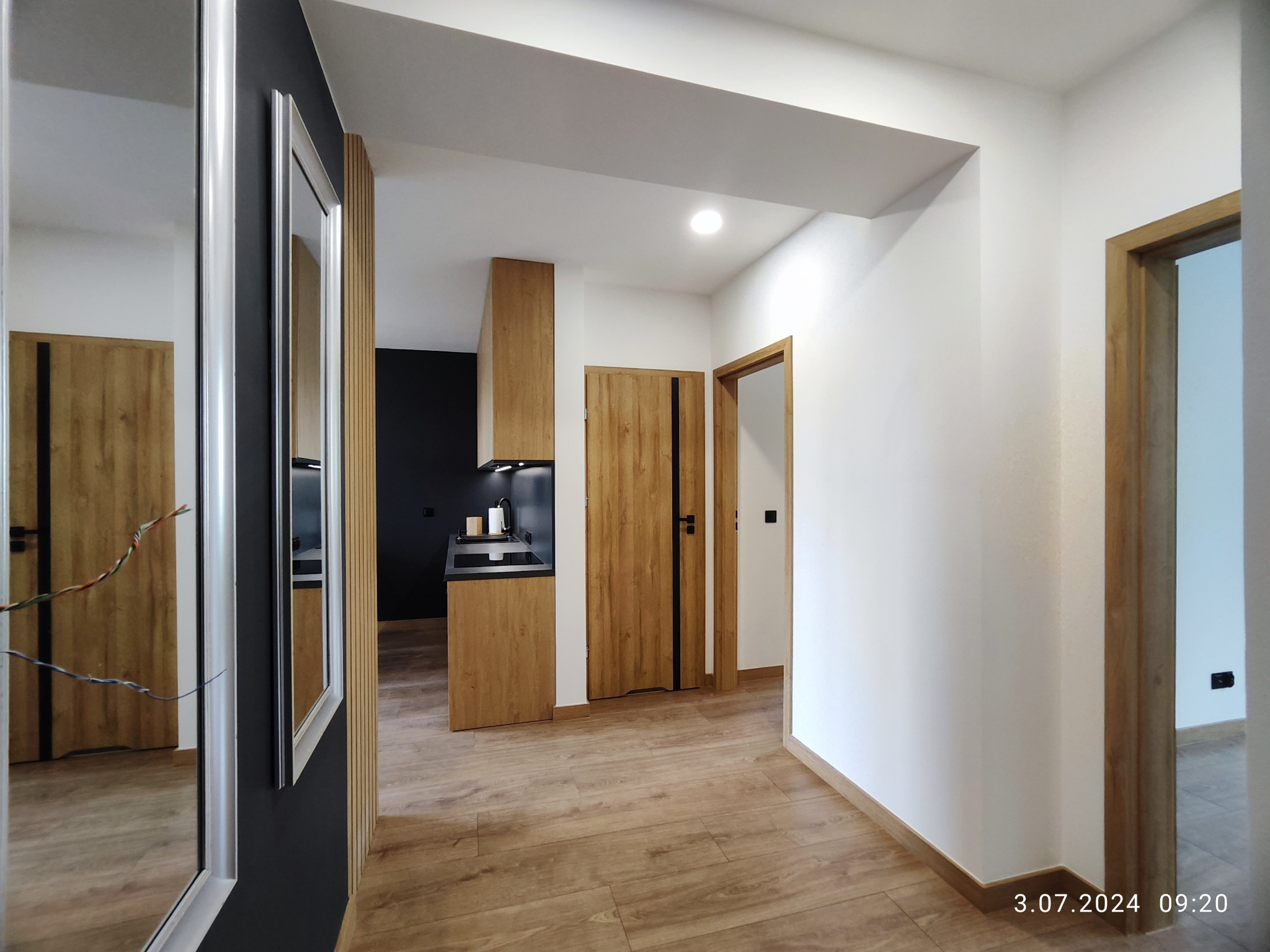 Nowoczesny korytarz z drewnianą podłogą, lustrem w srebrnej ramie i widokiem na kuchnię z czarną ścianą. Drzwi w kolorze drewna z czarnymi akcentami.