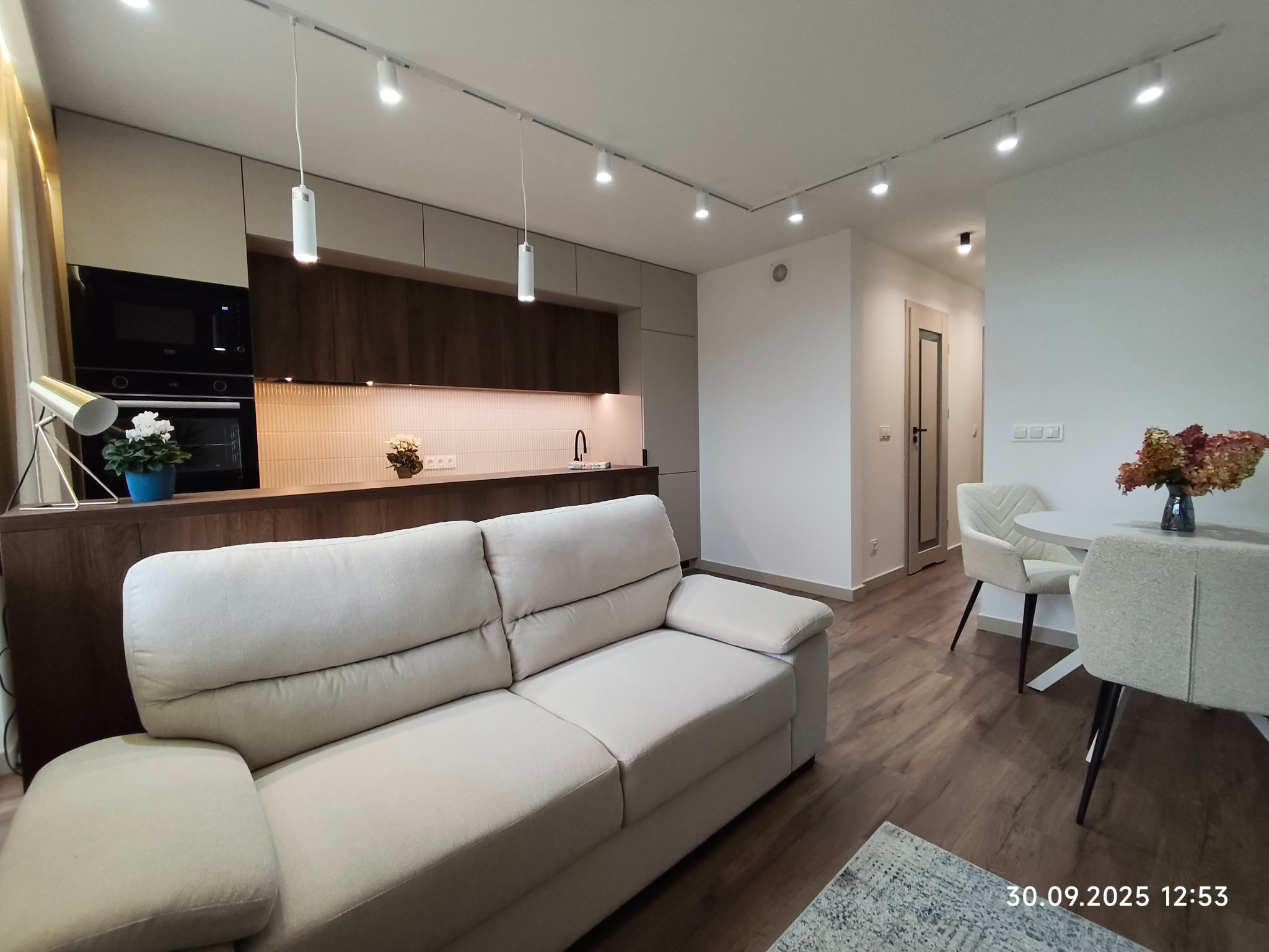 Nowoczesne wnętrze: beżowa sofa w salonie z aneksem kuchennym, drewniane podłogi, oświetlenie punktowe i minimalistyczny wystrój. Widok na stół z krzesłami.