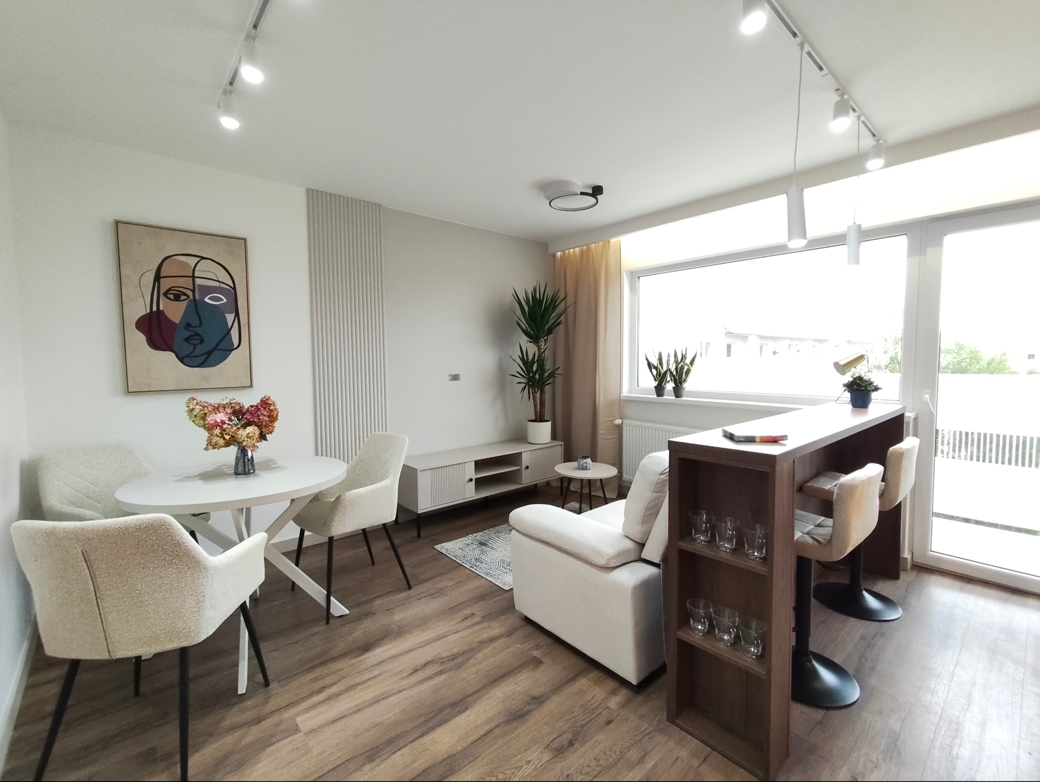 Jasne, nowocześnie urządzone wnętrze salonu w Kaletniku z drewnianą podłogą, białymi meblami i abstrakcyjnym obrazem na ścianie. Widoczne oświetlenie szynowe.