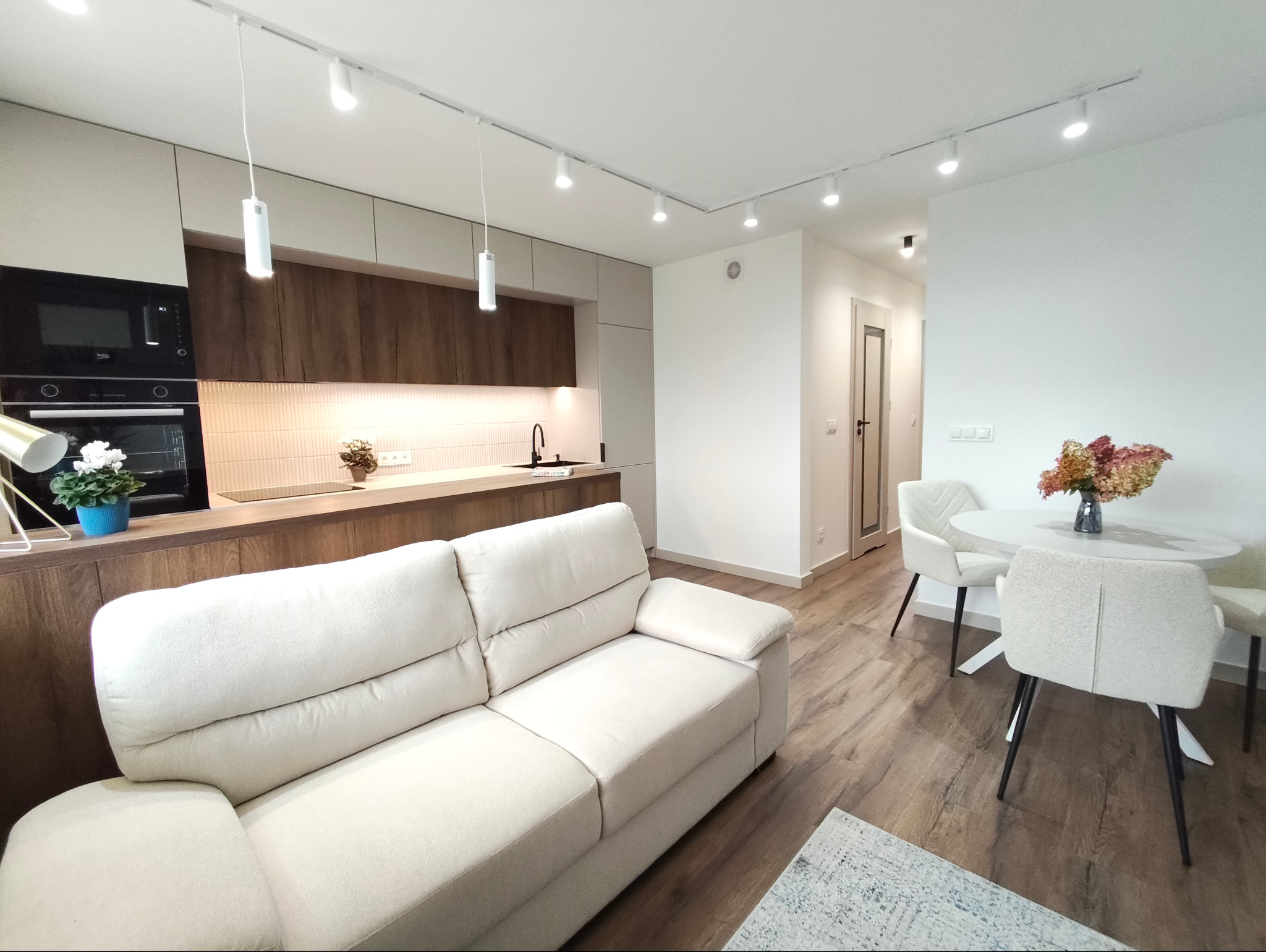 Jasne, nowocześnie urządzone wnętrze mieszkania z drewnianą zabudową kuchenną, białym narożnikiem i okrągłym stołem z krzesłami. Oświetlenie szynowe.