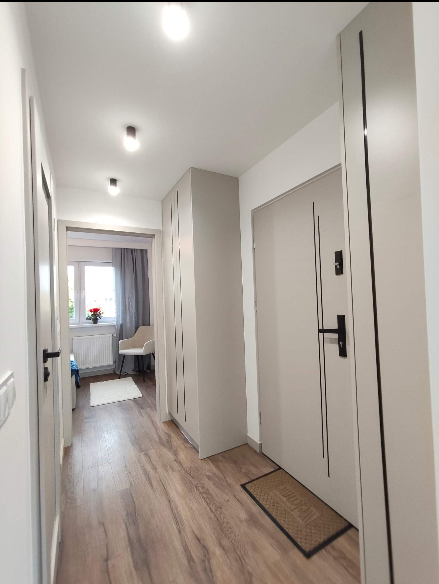 Jasny korytarz po remoncie: drewnopodobna podłoga, białe ściany, drzwi z czarnymi akcentami, szafa wnękowa i widok na pokój z kwiatami na parapecie.