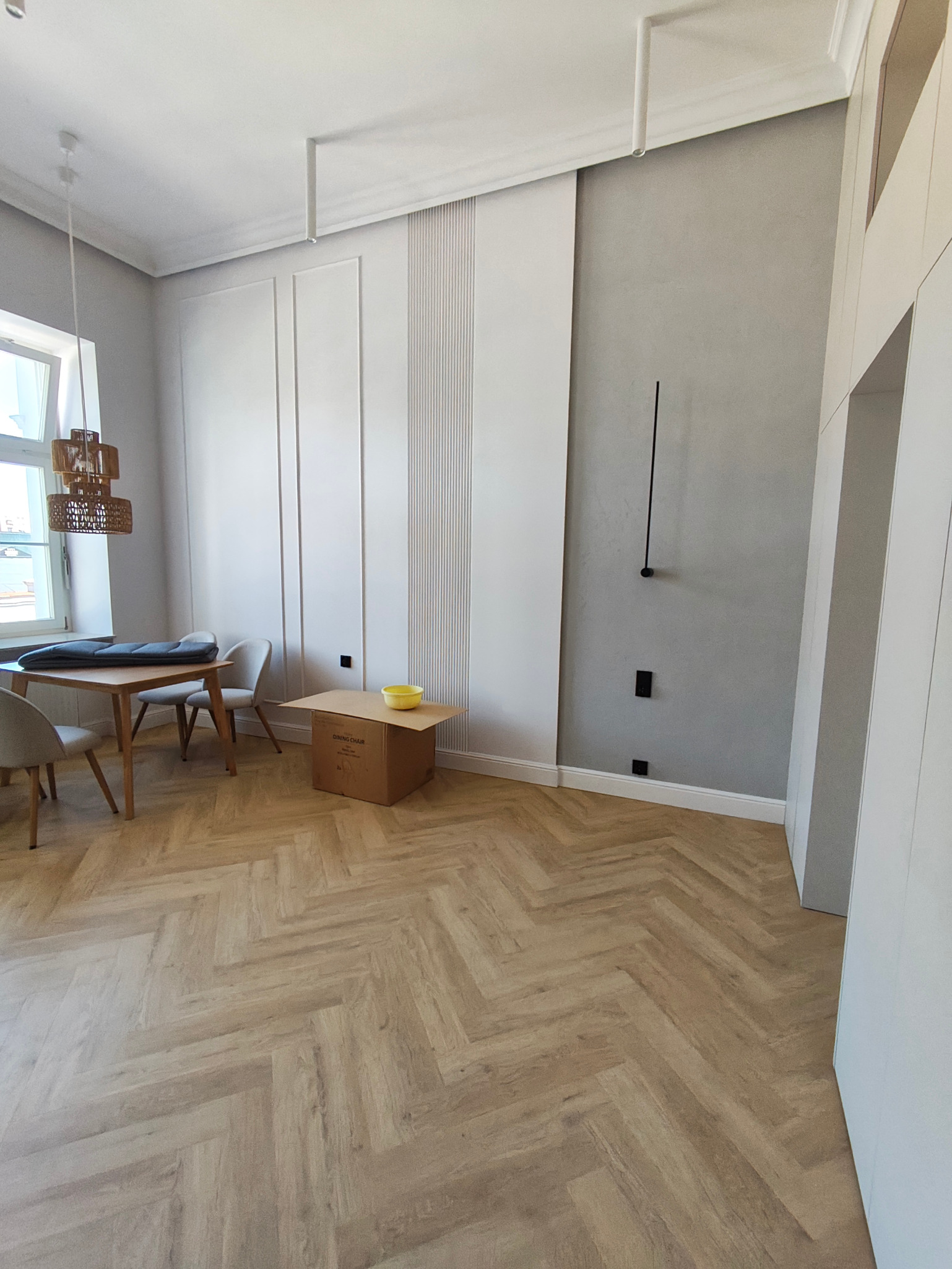 Jasne, przestronne wnętrze po remoncie z parkietem w jodełkę, dekoracyjnymi panelami ściennymi i minimalistycznym oświetleniem. Widoczne gniazdka elektryczne.