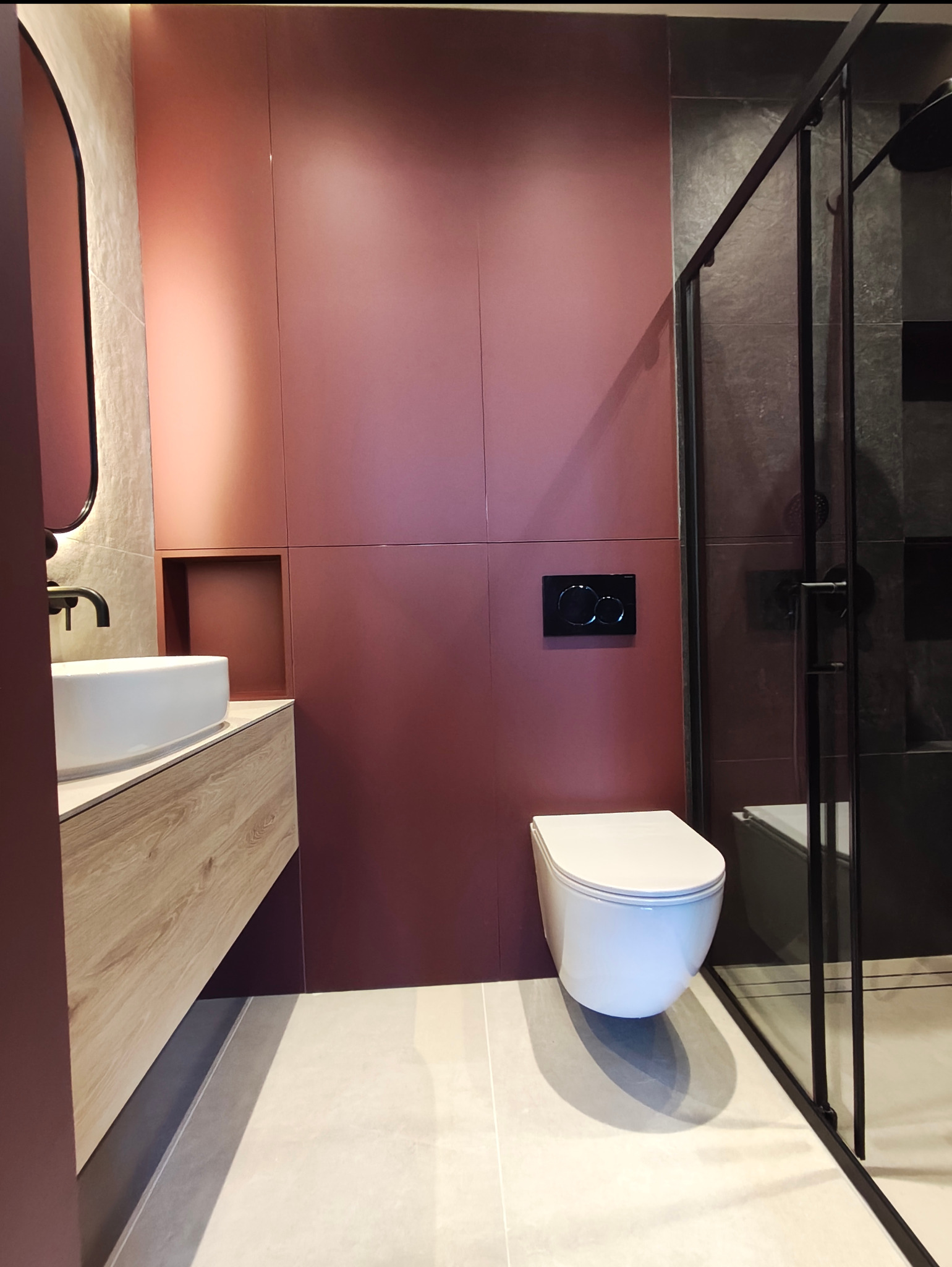 Nowoczesna łazienka z umywalką na drewnianej szafce, wiszącą toaletą i kabiną prysznicową z czarnymi ramami na tle ściany w kolorze bordo.