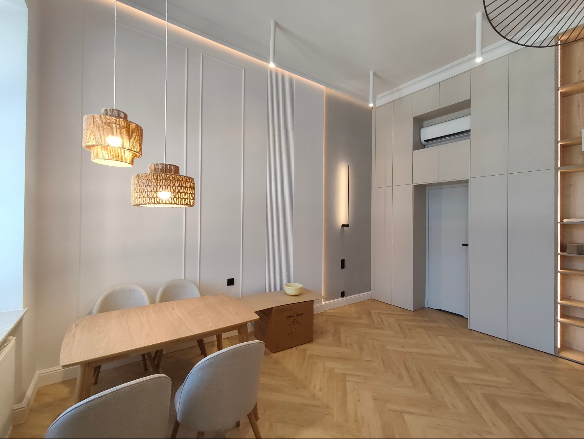 Jasne wnętrze jadalni z parkietem w jodełkę, drewnianym stołem i plecionymi lampami. Ściany zdobione sztukaterią i pionowym oświetleniem. Minimalistyczny design.