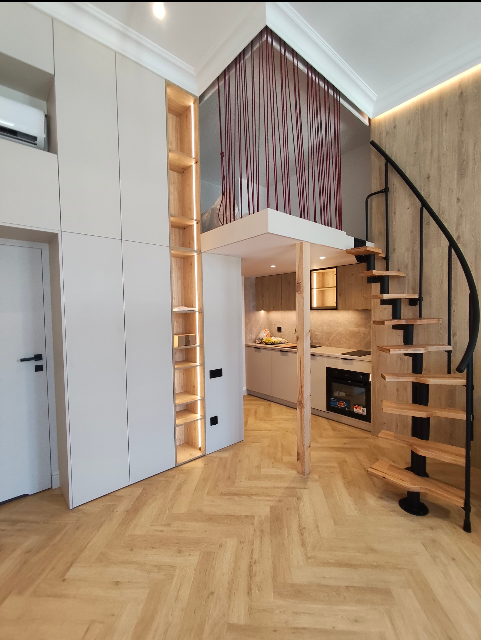 Nowoczesne wnętrze z antresolą: białe szafy, oświetlone półki, spiralne schody i parkiet w jodełkę. Widok na aneks kuchenny z drewnianymi akcentami.