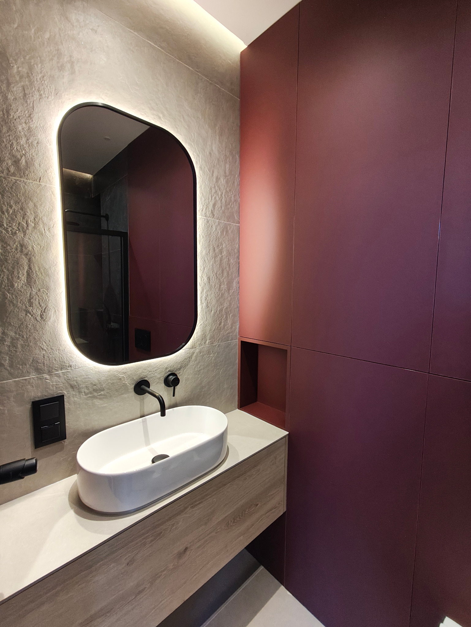 Nowoczesna łazienka z owalnym lustrem podświetlanym LED, białą umywalką nablatową i zabudową w kolorze burgund. Minimalistyczny design, szare płytki imitujące kamień.