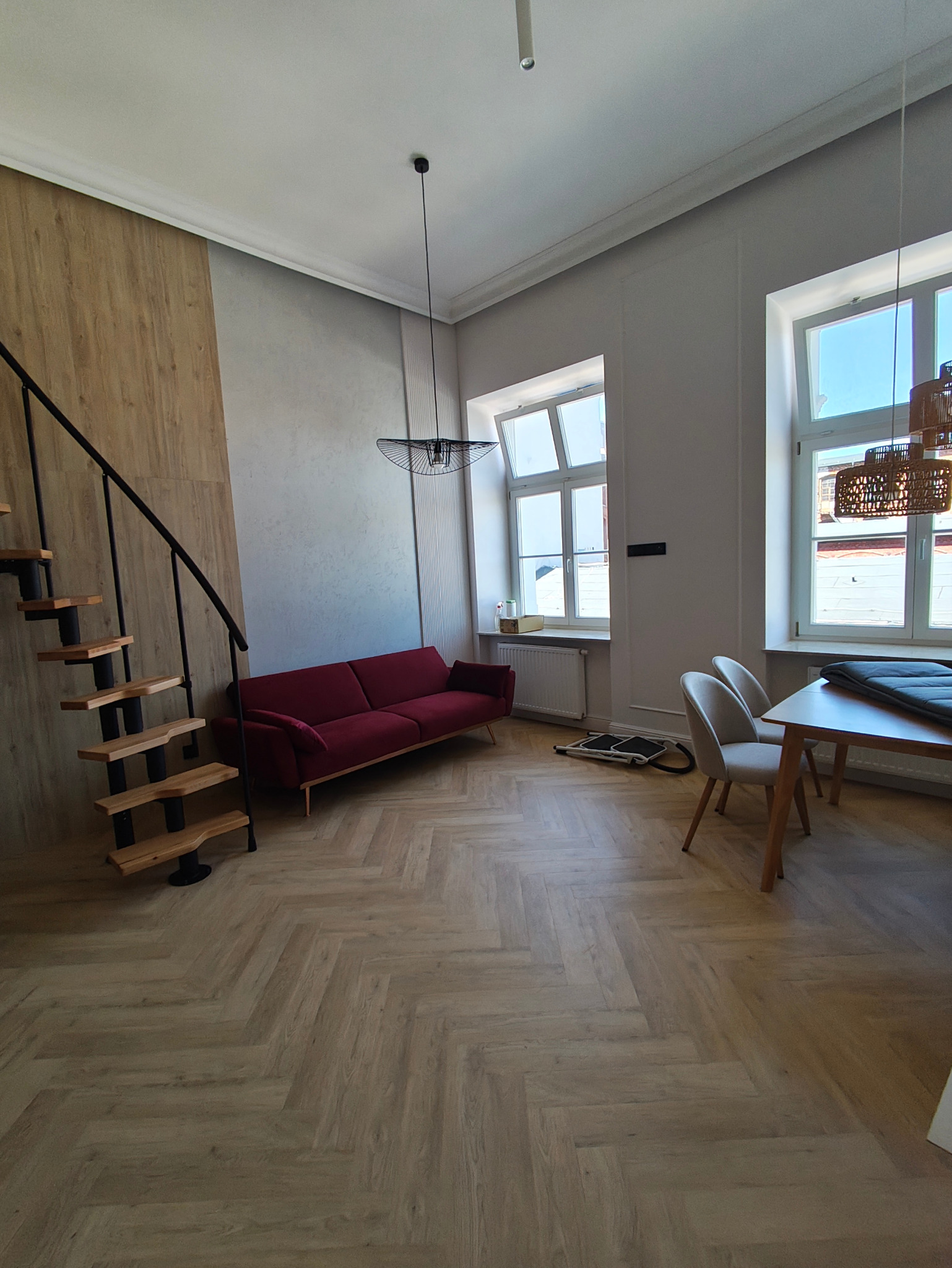 Jasne, przestronne wnętrze z parkietem w jodełkę, bordową sofą i schodami z drewnianymi stopniami. Dwa okna wpuszczają naturalne światło. Minimalistyczny wystrój.