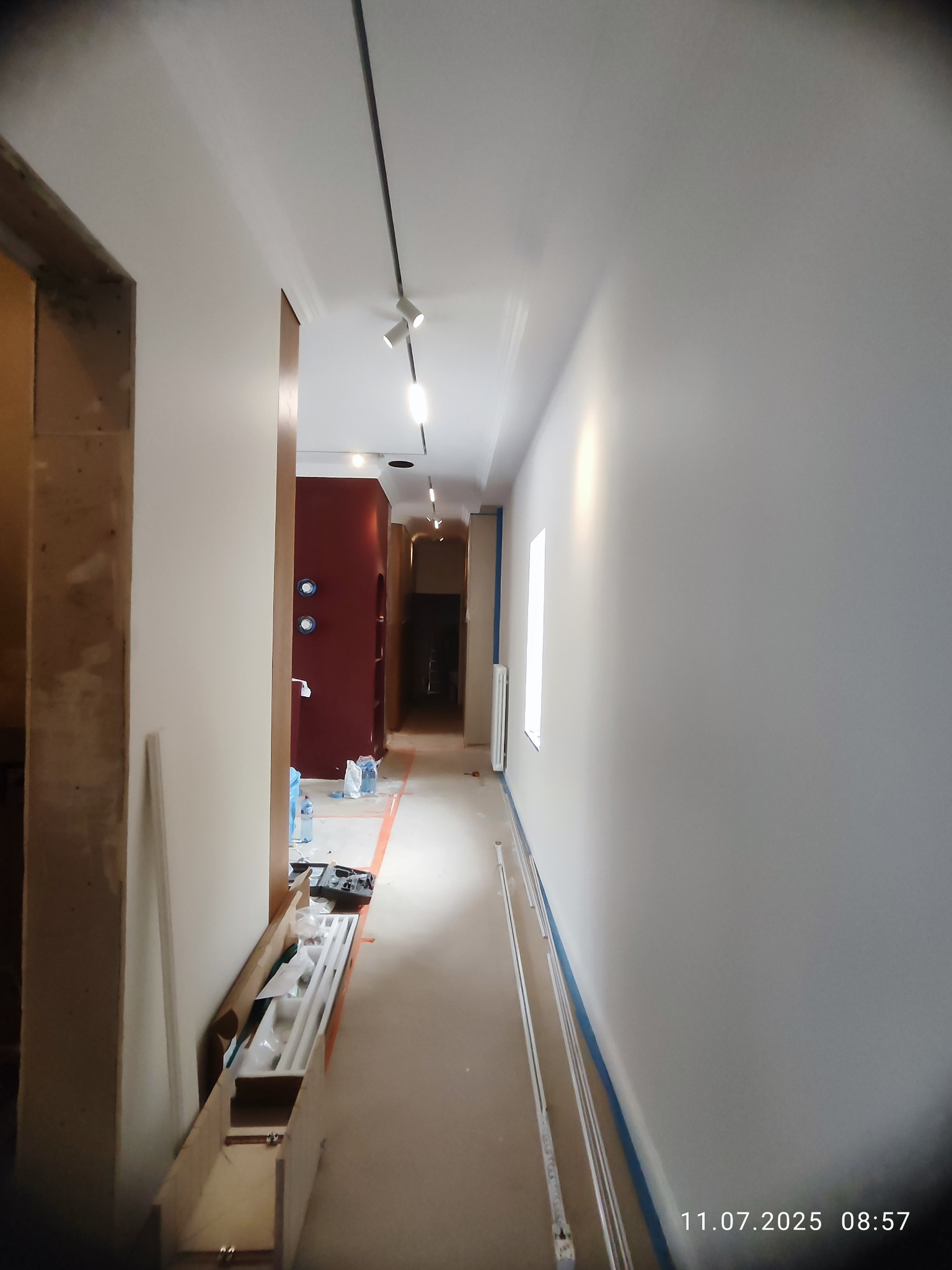 Korytarz w trakcie wykańczania: świeżo pomalowane ściany na biało, widoczne listwy przypodłogowe i oświetlenie szynowe. Prace wykończeniowe w toku.