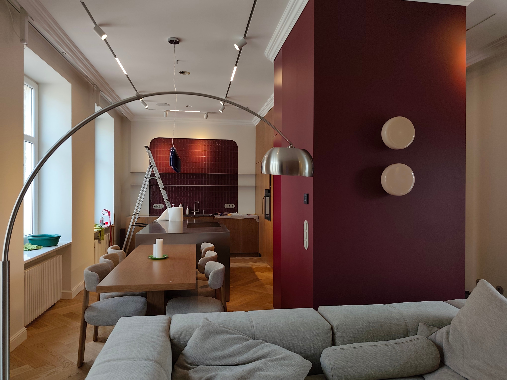 Nowoczesna kuchnia z burgundową ścianą, oświetleniem szynowym i łukową lampą. Widoczny stół z krzesłami i drabina malarska w tle. Parkiet w jodełkę.
