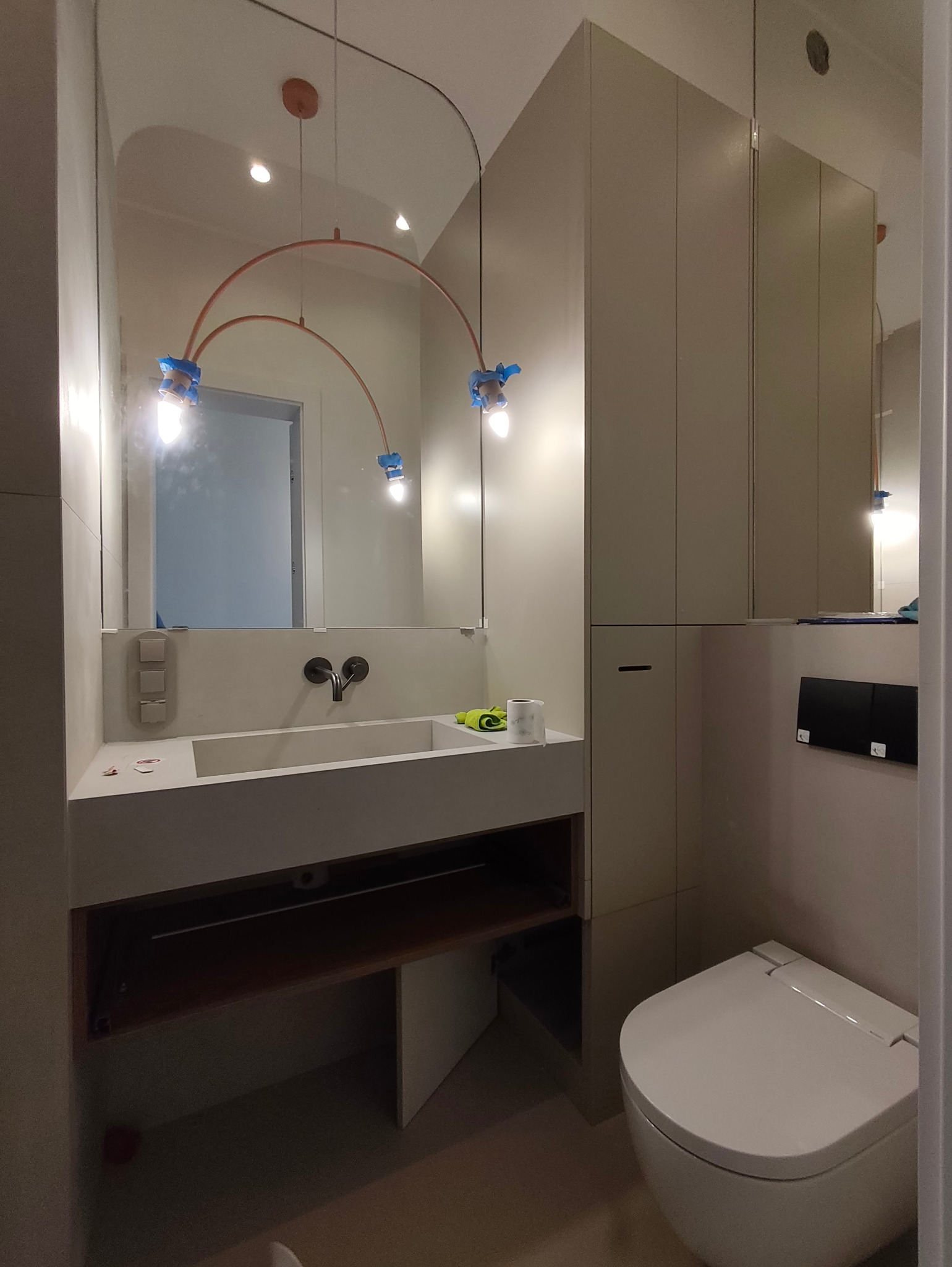 Nowoczesna łazienka z umywalką wpuszczaną w blat, wiszącą toaletą i oryginalnym oświetleniem. Widoczne zabezpieczenia na lampach sugerują etap wykończenia.