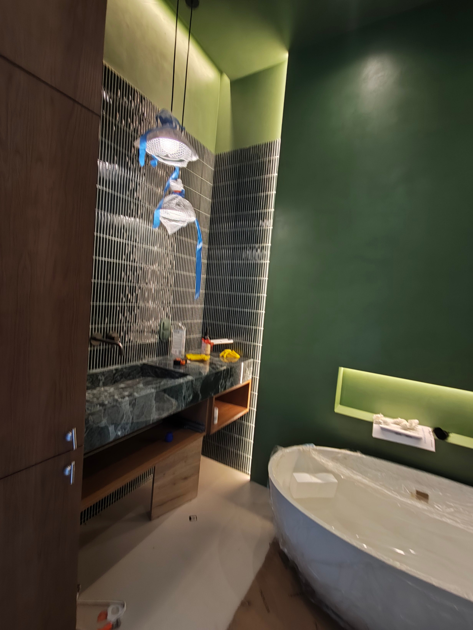 Nowoczesna łazienka w trakcie wykańczania: zielone ściany, mozaika, marmurowa umywalka i wolnostojąca wanna zabezpieczona folią. Widoczne narzędzia i lampy z niebieską folią.