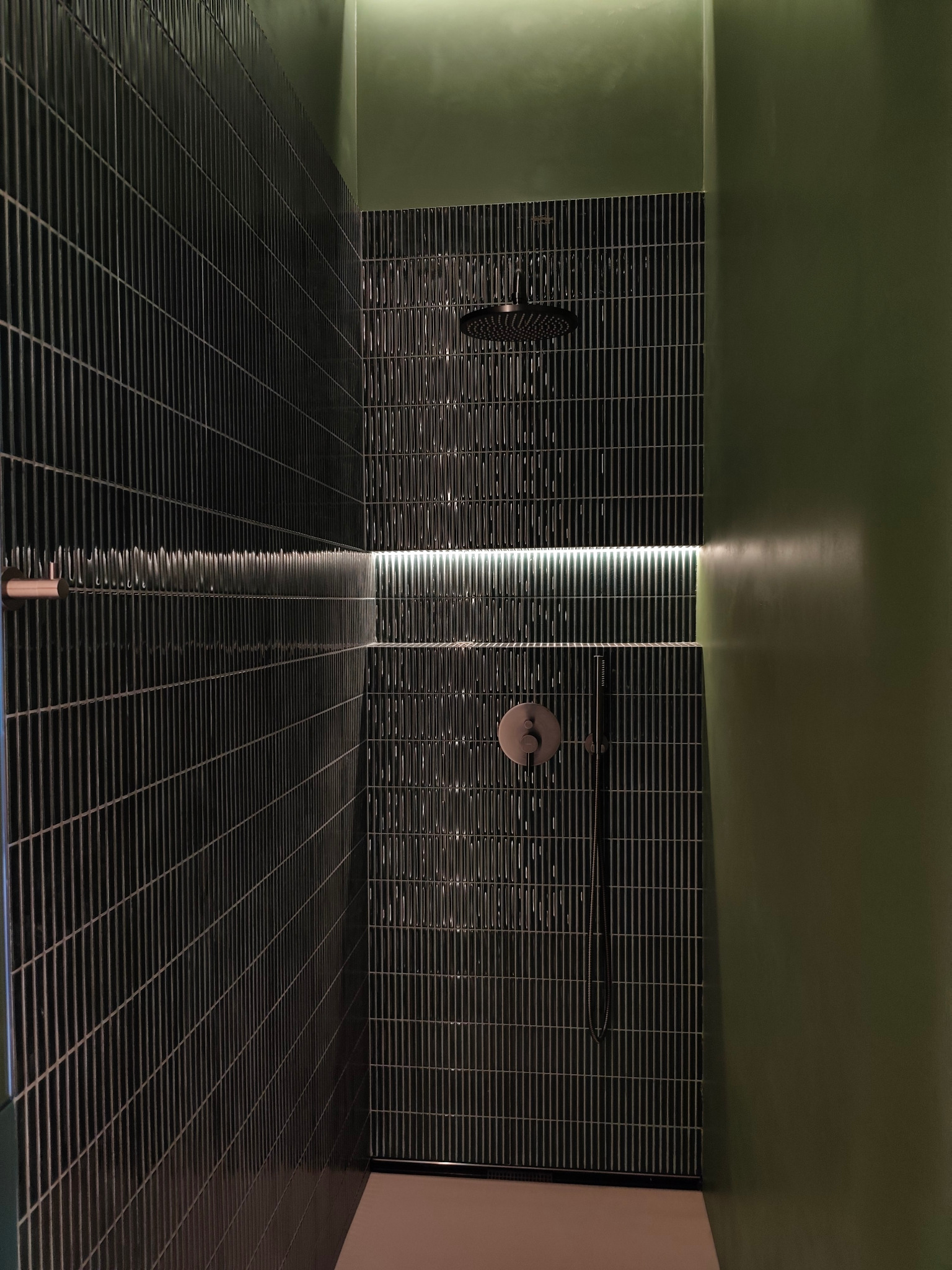 Nowoczesna kabina prysznicowa z ciemnymi, pionowo ułożonymi płytkami i subtelnym oświetleniem LED, kontrastująca z oliwkową ścianą. Minimalistyczny design.