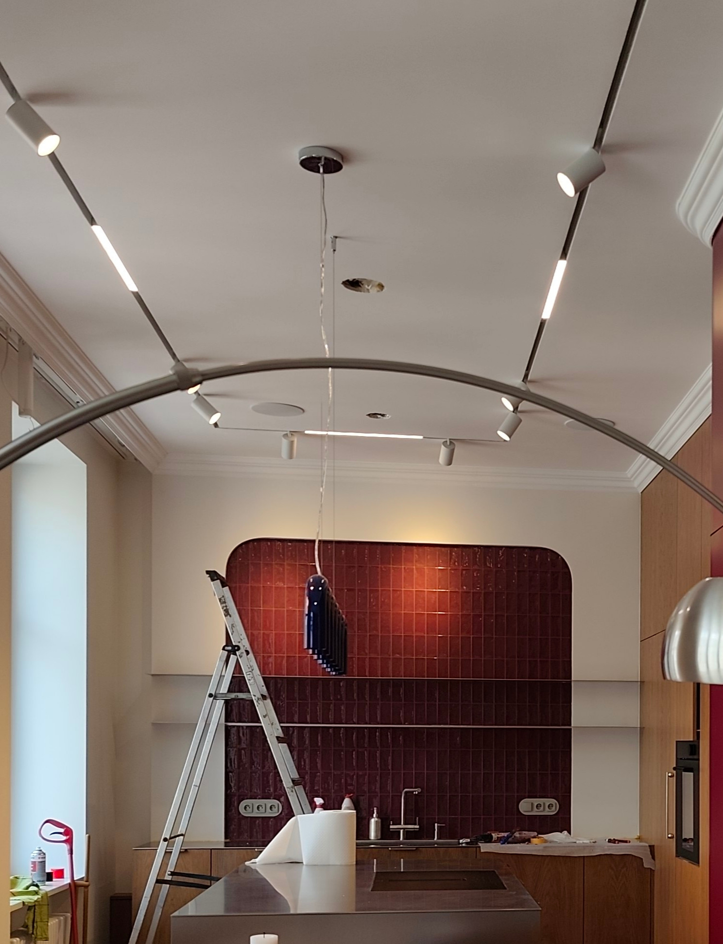 Kuchnia w trakcie montażu oświetlenia: szyna z reflektorami na suficie, drabina, bordowe płytki na ścianie. Widoczne elementy wyposażenia kuchni.