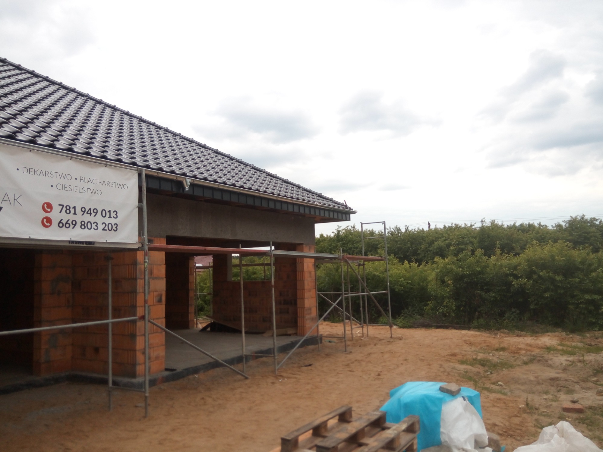 Budowa domu jednorodzinnego w trakcie prac dekarskich, widoczny nowy dach z dachówki ceramicznej, ściany z czerwonej cegły oraz rusztowania. Na pierwszym planie baner reklamowy firmy dekarskiej.