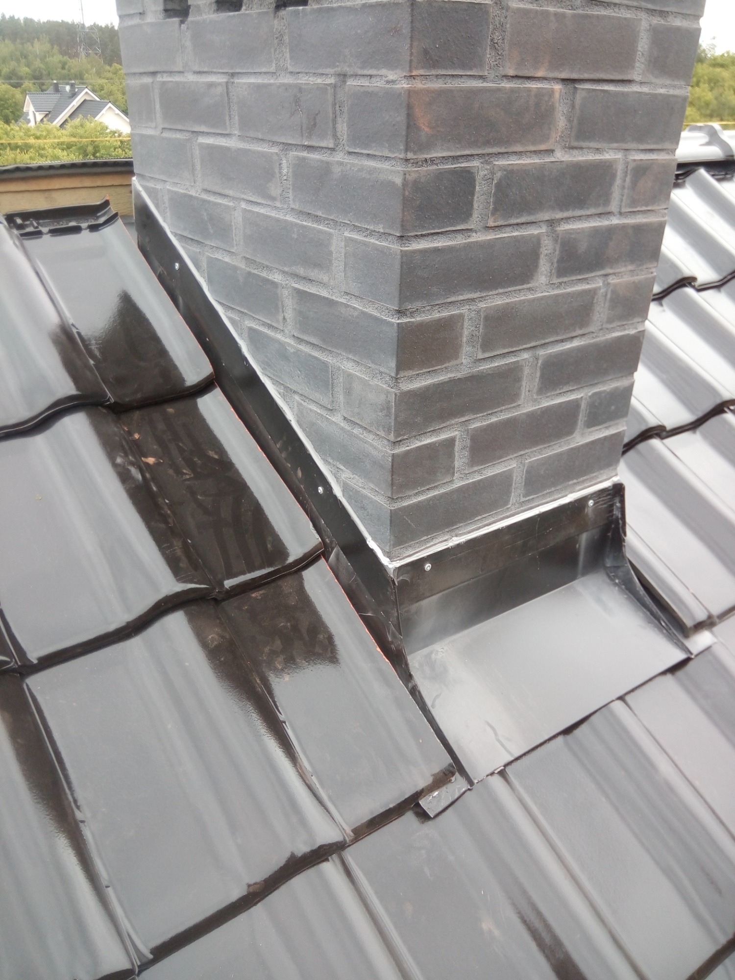 Błyszczące dachówki ceramiczne w kolorze grafitowym otaczają komin z ciemnej cegły, zabezpieczone obróbką blacharską z widocznymi nitami.