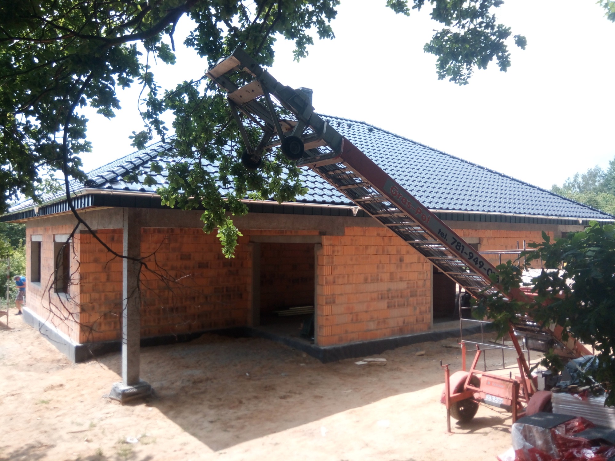 Nowy, ciemny dach z dachówki ceramicznej na budynku w trakcie budowy, z podnośnikiem do transportu materiałów na placu budowy, widoczne ściany z czerwonej cegły i elementy konstrukcyjne.